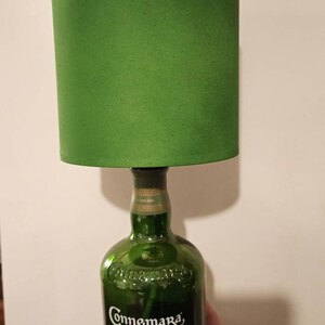 Pu&ograve; includere: Un paralume verde si trova sopra una bottiglia di vetro trasparente con un'etichetta verde su cui &egrave; scritto "Connemara Peated Single Malt Irish Whiskey Original" e "Clifden Co. Distilling".