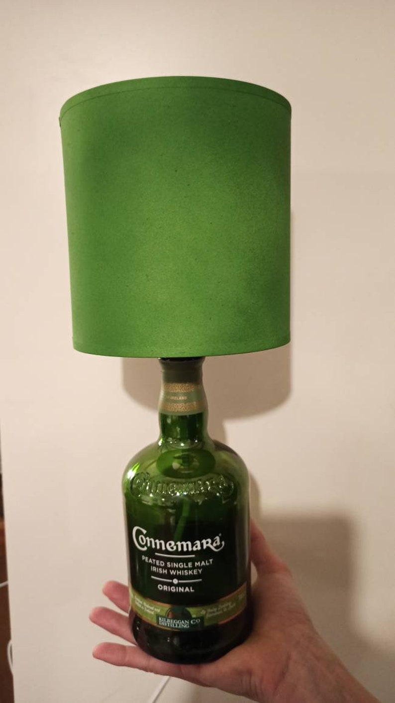 Pu&ograve; includere: Un paralume verde si trova sopra una bottiglia di vetro trasparente con un'etichetta verde su cui &egrave; scritto "Connemara Peated Single Malt Irish Whiskey Original" e "Clifden Co. Distilling".