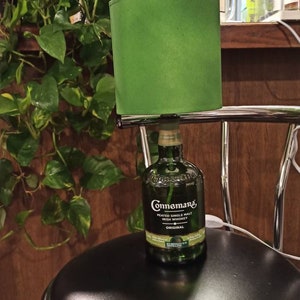 Pu&ograve; includere: Un paralume verde con una bottiglia di whisky irlandese Connemara Peated Single Malt al suo interno. La bottiglia &egrave; appoggiata su uno sgabello da bar nero con una base cromata.