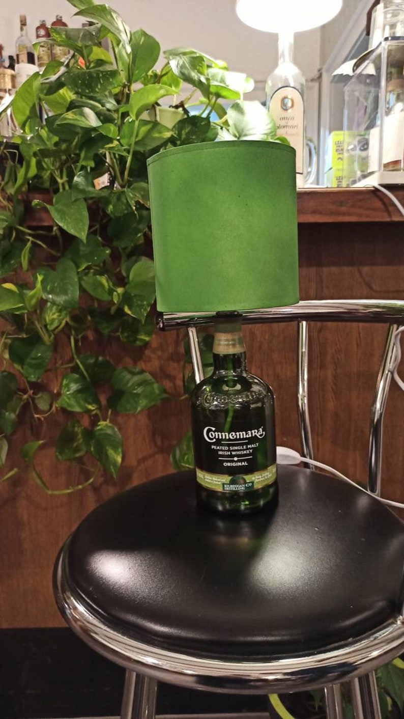Pu&ograve; includere: Un paralume verde con una bottiglia di whisky irlandese Connemara Peated Single Malt al suo interno. La bottiglia &egrave; appoggiata su uno sgabello da bar nero con una base cromata.