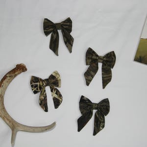 May include: Four camouflage fabric bows in various sizes, alongside a deer antler and a book titled "Good Luck" with an elk illustration. The bows have a brown and green pattern.
