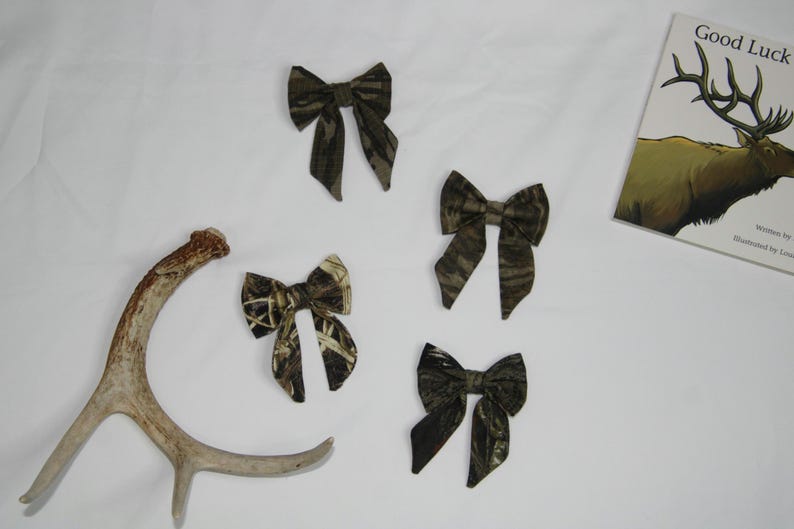 May include: Four camouflage fabric bows in various sizes, alongside a deer antler and a book titled "Good Luck" with an elk illustration. The bows have a brown and green pattern.
