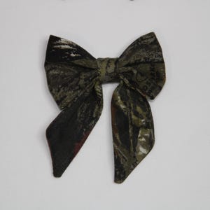 May include: A fabric bow tie with a camouflage pattern in shades of green, brown, and black. The bow tie is centered on a white background. The bow tie is a classic accessory.