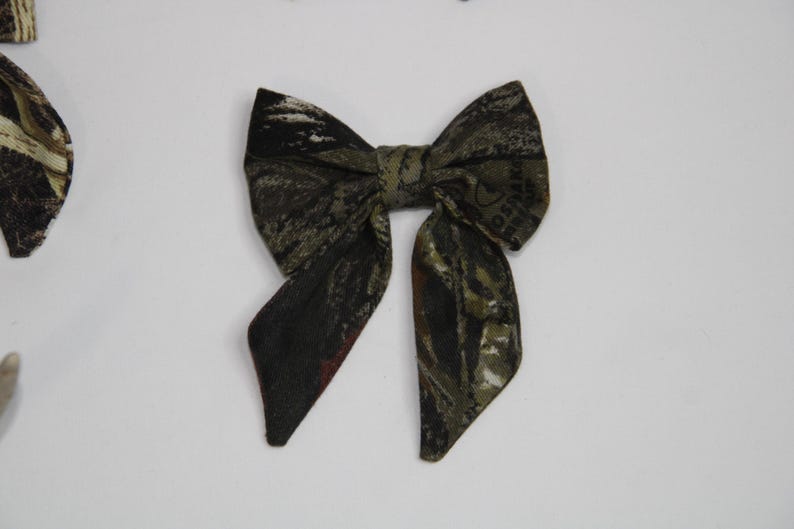 May include: A fabric bow tie with a camouflage pattern in shades of green, brown, and black. The bow tie is centered on a white background. The bow tie is a classic accessory.