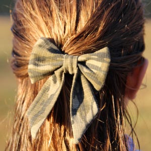 May include: A camouflage patterned bow hair accessory is secured in the hair. The bow is made of fabric with a green, brown, and black pattern. The hair is long and brown, with a slight wave. The background is blurred.