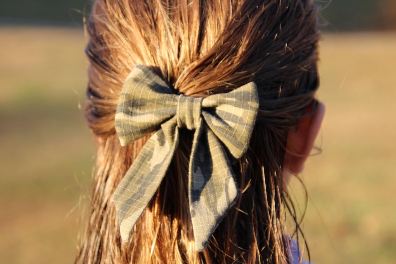 May include: A camouflage patterned bow hair accessory is secured in the hair. The bow is made of fabric with a green, brown, and black pattern. The hair is long and brown, with a slight wave. The background is blurred.