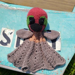 May include: A handmade crochet octopus doll with a red head, green accents, and a black beak. The octopus has gray arms and a gray skirt-like body. The doll is sitting on a weathered blue surface.
