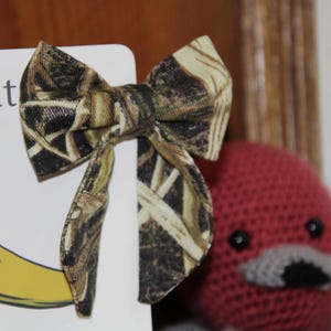 May include: A camouflage patterned bow tie is displayed next to a book with the word "Hunt" visible. The bow tie is made of fabric with a brown, green, and tan pattern. A red crocheted duck is in the background.