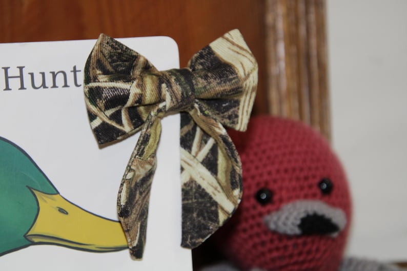 May include: A camouflage patterned bow tie is displayed next to a book with the word "Hunt" visible. The bow tie is made of fabric with a brown, green, and tan pattern. A red crocheted duck is in the background.