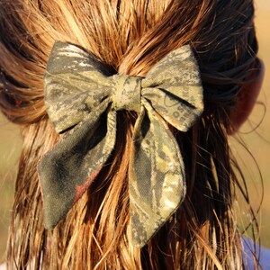 May include: A camouflage print hair bow, tied in a bow shape, is secured in the hair. The bow features shades of green, brown, and black. The hair is long and light brown. The bow is a hair accessory.