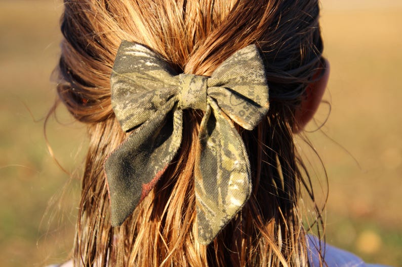 May include: A camouflage print hair bow, tied in a bow shape, is secured in the hair. The bow features shades of green, brown, and black. The hair is long and light brown. The bow is a hair accessory.