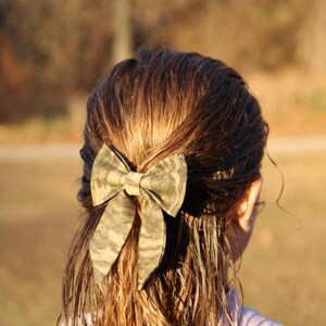 May include: A camouflage-patterned bow is secured in the hair. The bow is made of fabric with a green, brown, and tan design. The person's hair is wet and brown, with the bow positioned at the back of the head. The background is blurred.