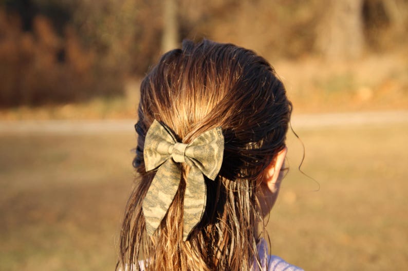May include: A camouflage-patterned bow is secured in the hair. The bow is made of fabric with a green, brown, and tan design. The person's hair is wet and brown, with the bow positioned at the back of the head. The background is blurred.