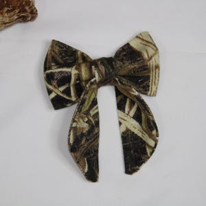 May include: A camouflage bow tie with a pattern of brown, green, and tan. The bow tie is made of fabric and has a classic bow shape with two long, pointed ends. The bow tie is set against a white background.