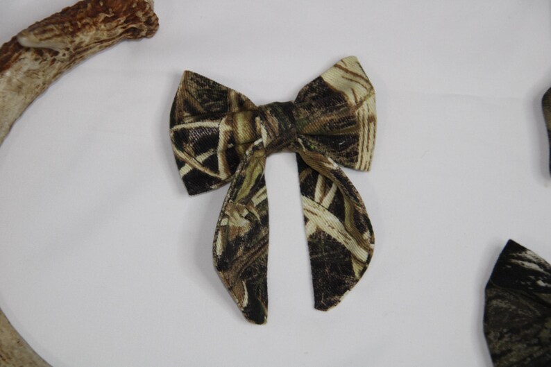 May include: A camouflage bow tie with a pattern of brown, green, and tan. The bow tie is made of fabric and has a classic bow shape with two long, pointed ends. The bow tie is set against a white background.