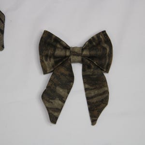 May include: A fabric bow tie with a camouflage pattern in shades of brown, tan, and green. The bow tie is centered in the image, with other bow tie pieces visible in the background. The bow tie has a classic shape.