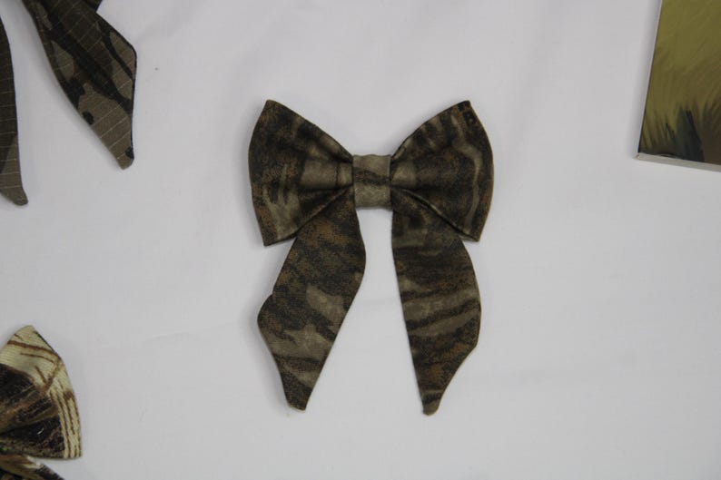 May include: A fabric bow tie with a camouflage pattern in shades of brown, tan, and green. The bow tie is centered in the image, with other bow tie pieces visible in the background. The bow tie has a classic shape.