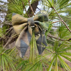 May include: A camouflage bow tie in shades of green, brown, and gray, set against a backdrop of pine needles. The bow tie is made of fabric with a woodland pattern, perfect for outdoor enthusiasts.