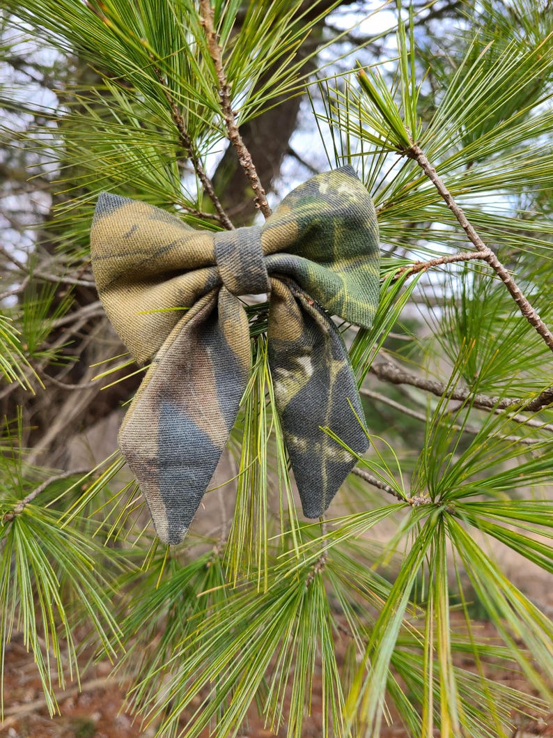 May include: A camouflage bow tie in shades of green, brown, and gray, set against a backdrop of pine needles. The bow tie is made of fabric with a woodland pattern, perfect for outdoor enthusiasts.