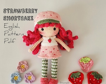 Puede incluir: Una muñeca de ganchillo hecha a mano que se asemeja a Strawberry Shortcake, con un vestido rosa, sombrero y cabello de hilo rojo. La muñeca tiene piernas a rayas verdes y blancas y zapatos rojos. También hay fresas y flores de ganchillo.
