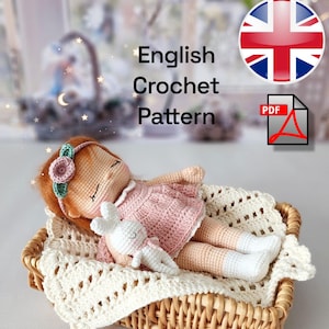 May include: A handmade crochet doll with red hair, wearing a pink dress, resting in a wicker basket on a cream-coloured crocheted blanket. A small white crochet bunny is next to the doll. The text "English Crochet Pattern" is visible.