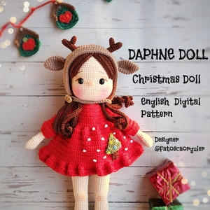 以下が含まれることがあります： 赤いドレスを着た、白いクリスマスツリーのデザイン、茶色のトナカイの帽子、緑と白のストライプの靴下を履いた、かぎ針編みのクリスマス人形。人形は木製の表面に立っており、背景には2つの包まれたプレゼントがあります。画像には「Daphne Doll Christmas Doll English Digital Pattern Designer @Fatoscaorguler」というテキストが表示されています。
