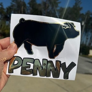 May include: A black pig sticker with gold trim and the name "PENNY" in reflective lettering. The sticker is held up against a blurred outdoor background, showcasing the pig's silhouette.