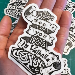 May include: Black and white sticker with a stylized image of a hammer and a scroll with the text "See you in Valhalla".