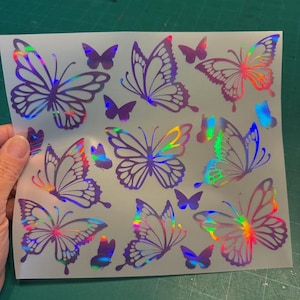 May include: A sheet of holographic butterfly stickers in various sizes. The butterflies have purple outlines and iridescent wings that reflect multiple colors. Some butterflies are solid purple.