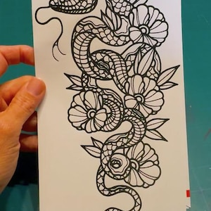 May include: A black and white line art illustration printed on a rectangular white sheet. The design features a winding snake with intricate scale patterns intertwined with several large, blooming flowers and pointed leaves in a vertical arrangement.