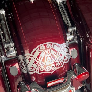 May include: Close-up of a burgundy motorcycle fender with a silver and white Norse-inspired graphic. The fender has chrome accents, red lights, and a license plate. The motorcycle's seat is visible in the background.