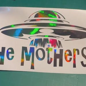 May include: A white sticker featuring a black UFO design with holographic accents. The text "The Mothership" is displayed below the UFO in a matching holographic font, creating a striking visual effect.