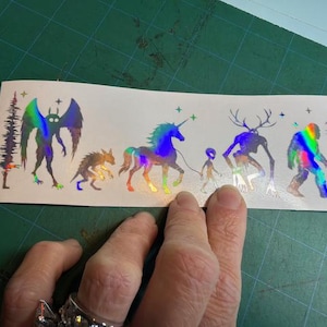 May include: A rectangular holographic sticker sheet featuring a line of mythical creatures including a moth-like figure, a small beast, a unicorn on a leash, a small alien, a deer-headed entity, and a sasquatch, all flanked by pine trees.