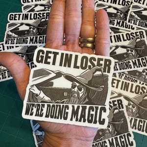 May include: A collection of black and white stickers with the text "GET IN LOSER" and "WE'RE DOING MAGIC". The stickers feature a wizard driving a car. The stickers are held in a hand.