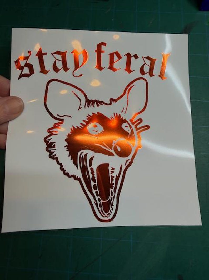 May include: A metallic orange vinyl decal on a white backing sheet featuring a graphic illustration of a snarling opossum head with its mouth wide open. Above the animal, the text "stay feral" is printed in a stylized, gothic-inspired font.