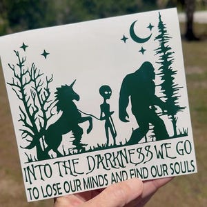 May include: A white square sticker with a dark green illustration of a unicorn, alien, and Bigfoot walking together. The text "INTO THE DARKNESS WE GO TO LOSE OUR MINDS AND FIND OUR SOULS" is below the illustration.