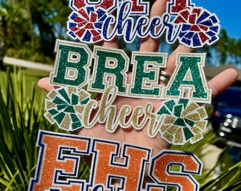 May include: Three glitter cheerleading spirit patches with the text "SFP cheer", "BREA cheer", and "EHS cheer" in white letters. Each patch features a glittery pom-pom in the school colors.
