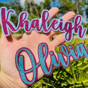 May include: Two personalized names, "Kaleigh" and "Olivia", are spelled out in sparkly glitter letters. The letters are pink and blue and are held in a hand.