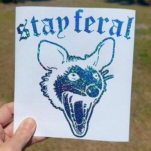 May include: A white square decal featuring a blue and green holographic glitter graphic of an opossum with its mouth open in a hiss. The text "stay feral" is printed above the opossum head in a gothic-style font with the same holographic finish.