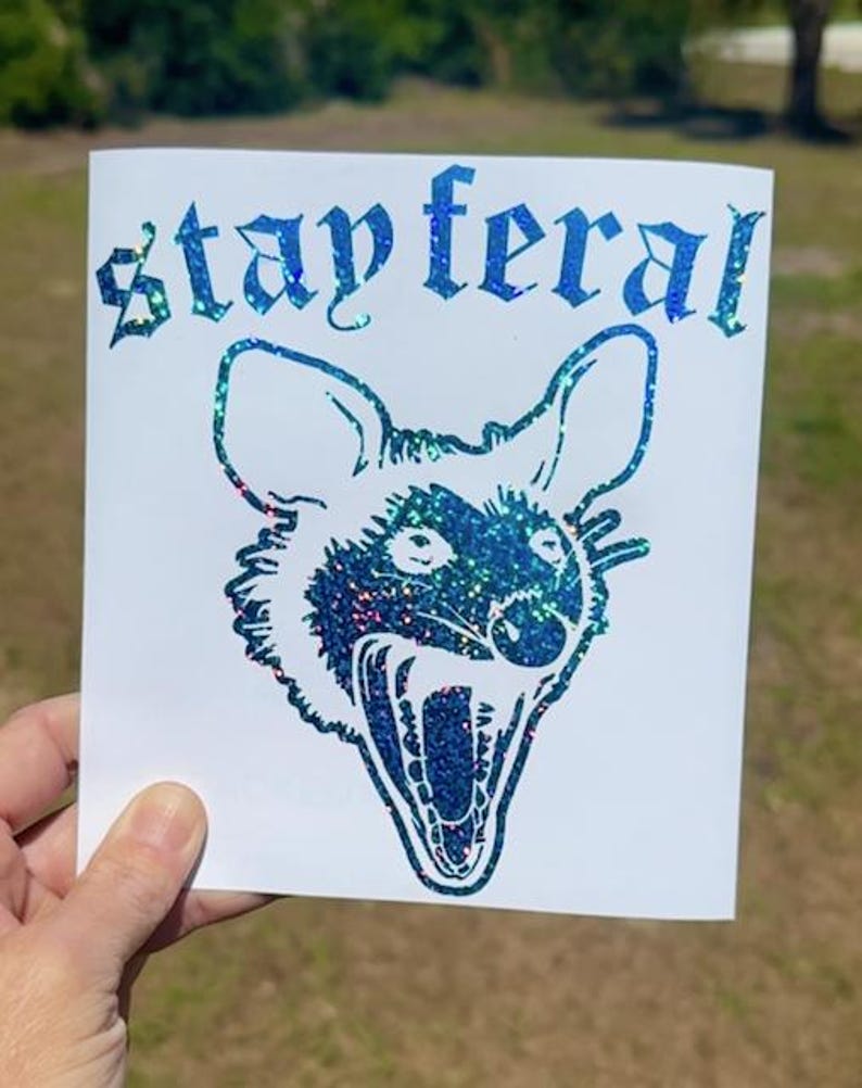 May include: A white square decal featuring a blue and green holographic glitter graphic of an opossum with its mouth open in a hiss. The text "stay feral" is printed above the opossum head in a gothic-style font with the same holographic finish.
