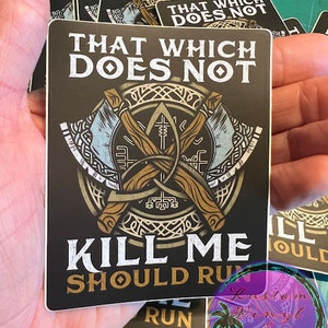 May include: A black and gold sticker with the text "That which does not kill me should run". The sticker features a stylized image of two crossed axes with a Celtic knot design.