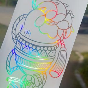 May include: A rainbow iridescent sticker with a detailed line drawing of a knot with a tassel and a decorative top.