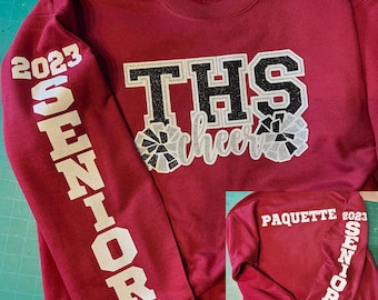 May include: Maroon sweatshirt with white lettering. The front features "THS cheer" with glitter accents. The left sleeve says "2023 SENIOR". A smaller image shows the back with "PAQUETTE 2023 SENIOR".