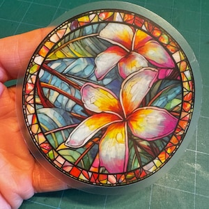May include: A round sticker with a stained glass design featuring two pink and yellow flowers with intricate details. The flowers are surrounded by green leaves and a colorful mosaic border.