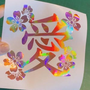 May include: A white sticker featuring the Japanese character for "love" in a holographic, rainbow color. The character is surrounded by holographic cherry blossom flowers, creating a vibrant and eye-catching design.