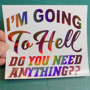 May include: A white sticker with the text "I'M GOING To Hell DO YOU NEED ANYTHING??" in a holographic, rainbow-colored font. The sticker is held against a green cutting mat.