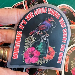 May include: A black and white sticker with a colorful bird, a grenade, and flowers. The sticker says "Sometimes it's the little things that count the most."