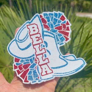 May include: A white glitter cheer boot with a red and blue pom-pom and the name "BELLA" in red glitter.