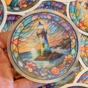 May include: A round sticker with a stained glass design featuring a lighthouse on a rocky shore with a sunset in the background. The lighthouse is white with a blue roof and a blue and white striped tower. The sticker has a silver border.