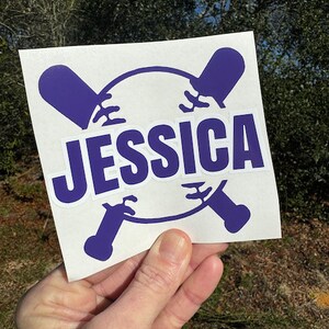 Op de afbeelding: Een witte sticker met een paars honkbal- en knuppelontwerp. De naam "JESSICA" is in paarse letters geschreven.